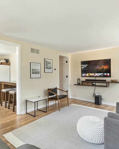 Open-plan living room with gray sofa, wall-mounted TV, wooden dining table, and kitchen visible through doorway