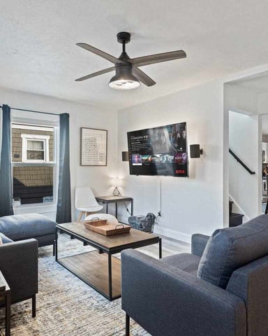 Open-plan living room with dark gray armchairs, ceiling fan, wall-mounted TV, and stone accent wall leading to dining area