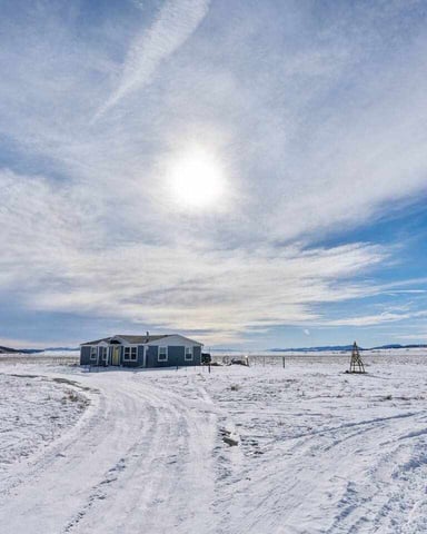 Dark green house on snowy prairie with dirt road, distant mountains and blue sky with sun