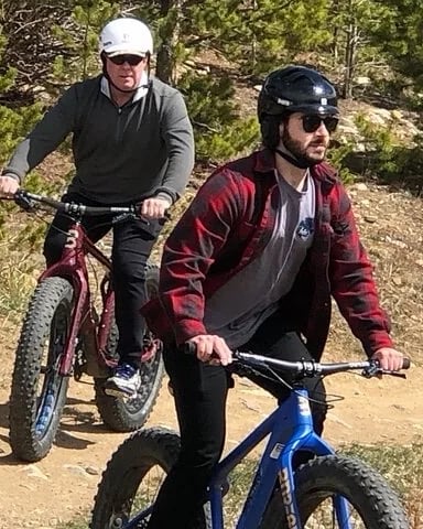 Two mountain bikers riding on a dirt trail through a forested area with pine trees