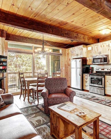 Open-plan cabin interior with wood beams, kitchen, fireplace, and seating area overlooking deck