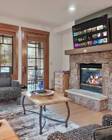 Living room with stone fireplace, gas insert, flat-screen TV, patterned armchair, wood coffee table, and balcony access with mountain views.