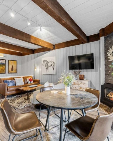 Open-plan living room with exposed wood beams, round dining table, leather sofa, fireplace, and white shiplap walls