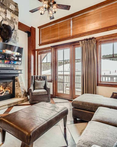 Luxury bedroom with stone fireplace, flat-screen TV, wood beams, and glass doors opening to snowy mountain view