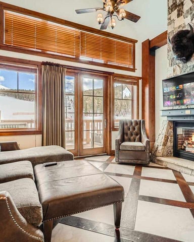 Living room with stone fireplace, wood-framed glass doors opening to snowy mountain view, leather seating, and ceiling fan.