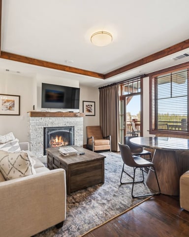 Spacious living room with fireplace, TV, beige seating, wood beams, and expansive windows overlooking landscape