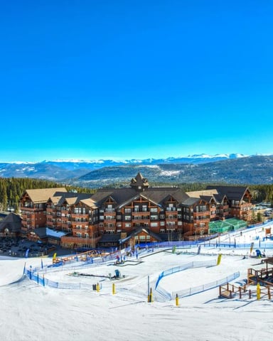 Ski resort village with red-brick lodges, snow-covered slopes, and snow-capped mountains in the distance under clear blue sky.