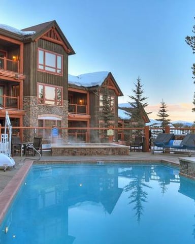 Mountain resort with heated outdoor pool, stone and wood lodge buildings, pine trees, and snow-capped peaks at dusk