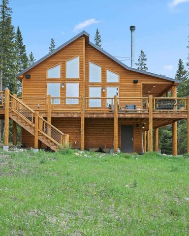 Modern log cabin with large windows and wraparound deck surrounded by tall pine trees and grassy clearing