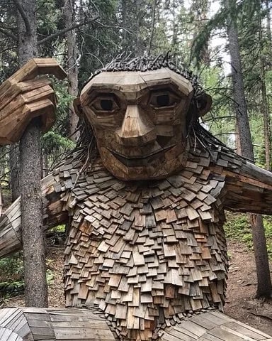 Large wooden troll sculpture with shingle-textured body and raised arm in forest surrounded by tall trees