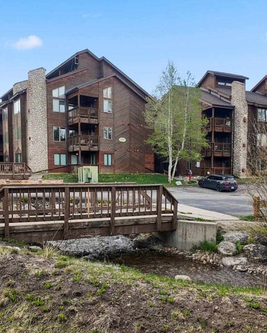 Multi-story mountain lodge buildings with stone chimneys and wooden decks, wooden bridge over stream in foreground, surrounded by evergreen trees and aspen.