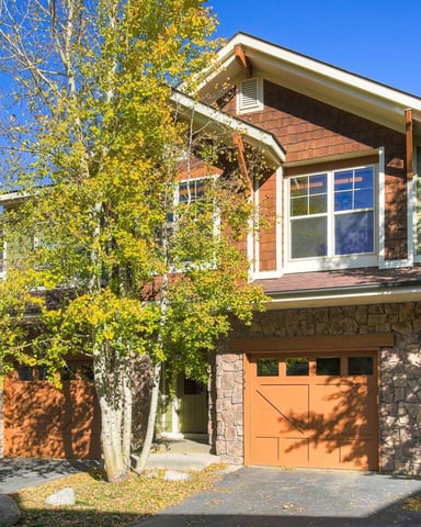 Modern brick and stone house with orange garage doors, large windows, and fall foliage surrounding the driveway.