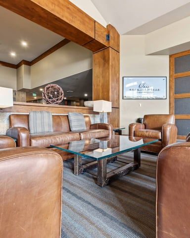 Modern living room with brown leather seating, glass coffee table, wood beams, and open kitchen with stone backsplash