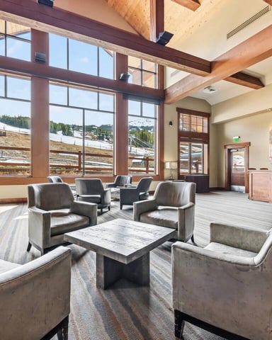 Spacious lodge lobby with exposed wood beams, large windows overlooking snowy mountains, and upholstered seating arranged around a central table.