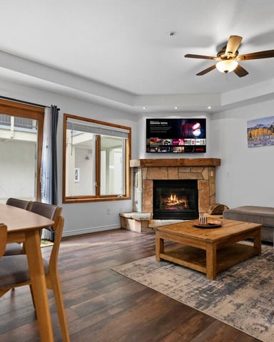 Open-plan living room with stone fireplace, gray sofa, wooden dining table, and TV mounted above mantel