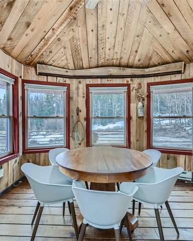 Rustic wood sunroom with red-framed windows, round wooden table, white chairs, and forest lake views