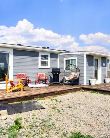 Modern mobile home with blue and white exterior, wooden deck with colorful chairs, grill, and hanging egg chair on rural property