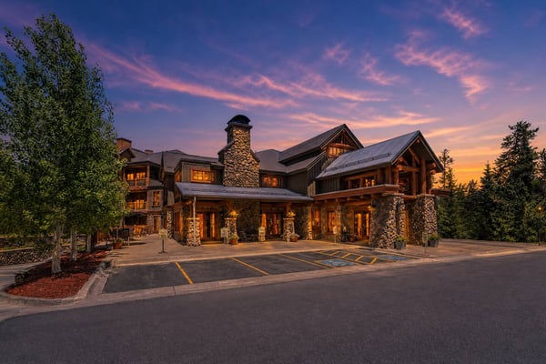 Large modern lodge with stone chimneys and timber accents at dusk, surrounded by evergreen trees and illuminated windows