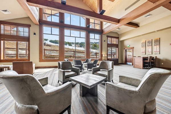 Spacious lodge lounge with exposed wood beams, floor-to-ceiling windows overlooking mountains, and seating areas with upholstered chairs around a stone table.