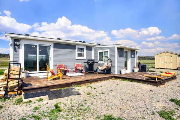 Modern mobile home with gray and white siding, wooden deck with colorful chairs, grill, and hanging egg chair on rural property