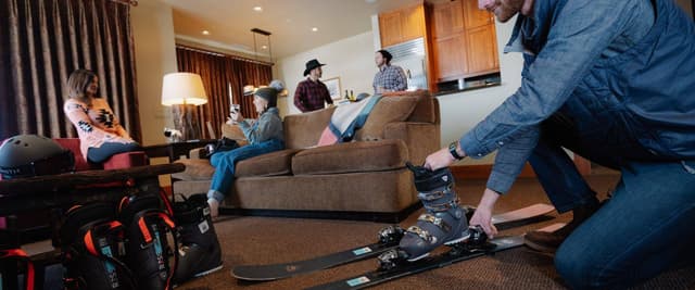 Man adjusting ski boots in open-plan living room with guests relaxing on couches and kitchen visible in background