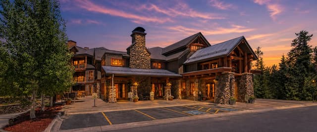 Large modern lodge with stone chimneys and timber accents at dusk, surrounded by evergreen trees and illuminated windows.