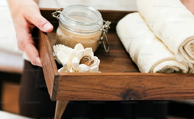 Wooden tray with rolled towels, glass jar, and white flower held by person's hand