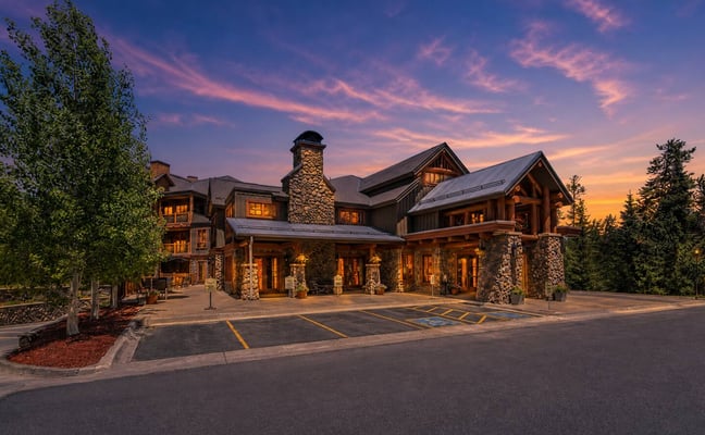Large modern lodge with stone chimneys and wood accents at dusk, surrounded by evergreen trees and illuminated windows