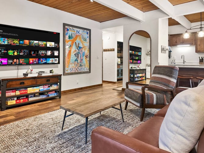 Open-plan living room with wood beams, leather sofa, mid-century chair, wood coffee table, Netflix-displaying TV, and kitchen visible in background