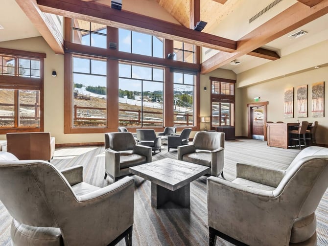 Spacious lodge lounge with exposed wood beams, floor-to-ceiling windows overlooking mountains, and seating areas with upholstered chairs around a stone coffee