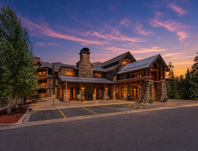 Large modern lodge with stone chimneys and timber accents at dusk, surrounded by evergreen trees and illuminated windows